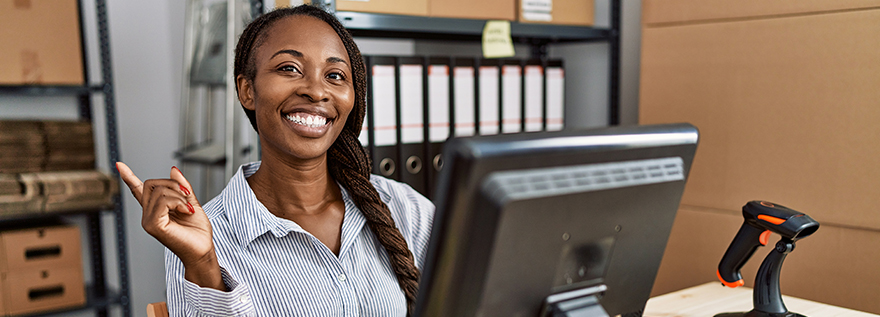 A Black business owner sits at a computer with inventory on shelves behind her. She is smiling and pointing back toward her inventory with her right hand.